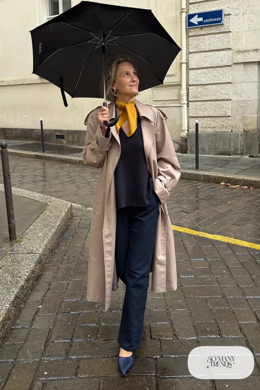 Petite woman in a belted short trench over a navy knit top and navy pants with a mustard scarf and flats, outfit 23