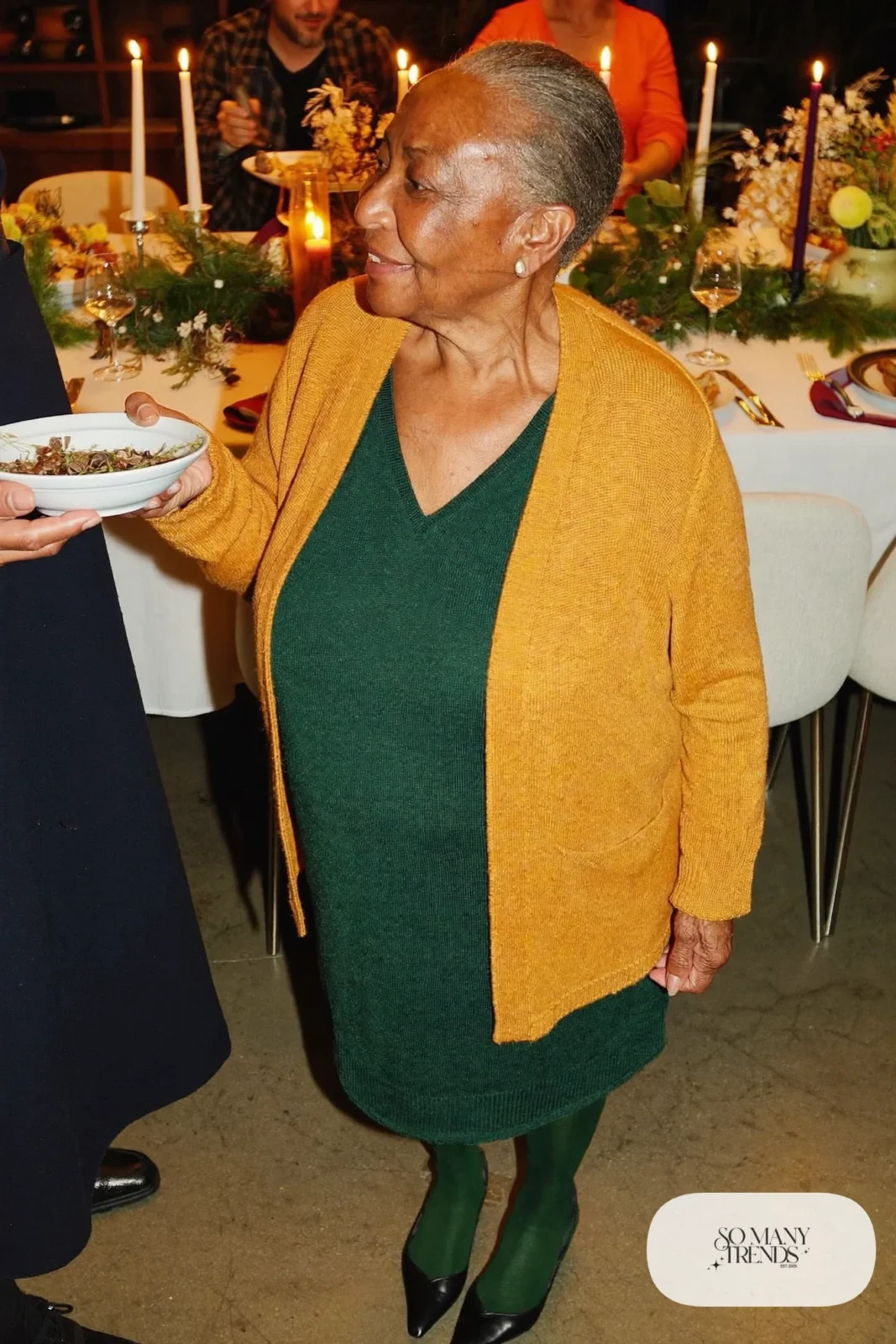 Petite woman in a hip-length mustard cardigan over a dark green midi dress with dark tights and pumps, outfit 28
