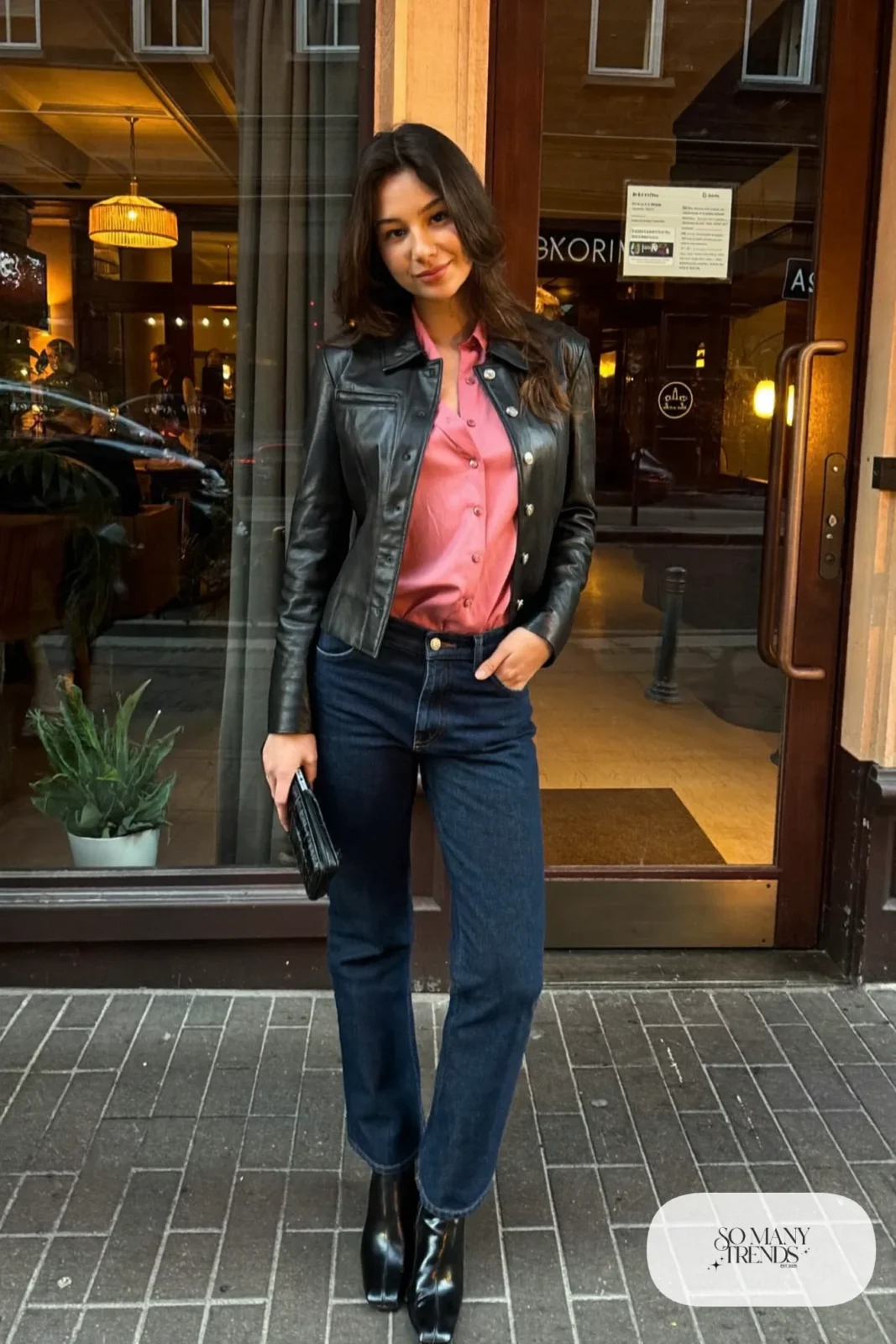 Woman in dark indigo jeans, dusty pink satin blouse, black leather jacket and block-heel boots, look 12