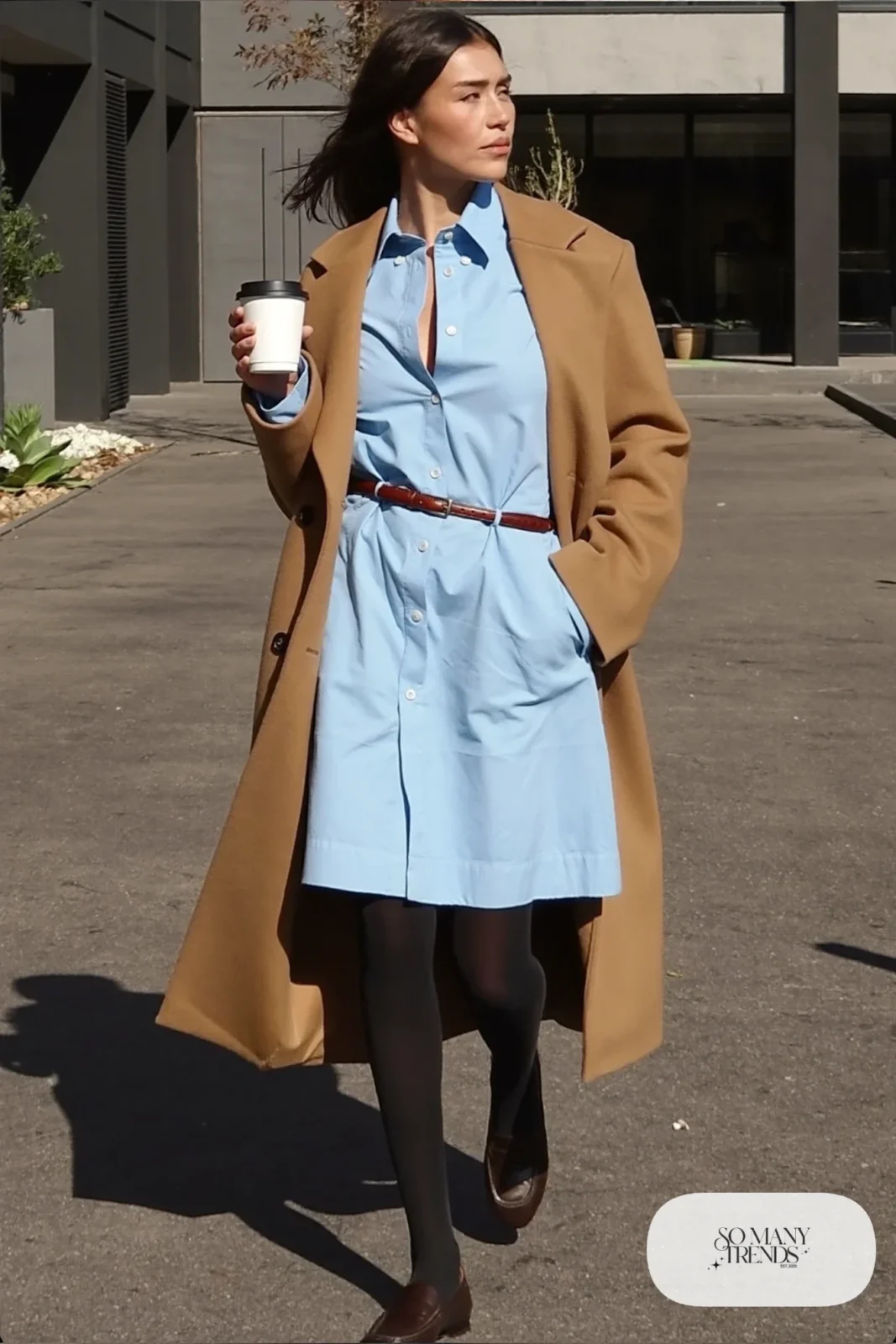 Woman in a sky blue belted shirt dress with a long camel coat, black tights and brown loafers, outfit 6