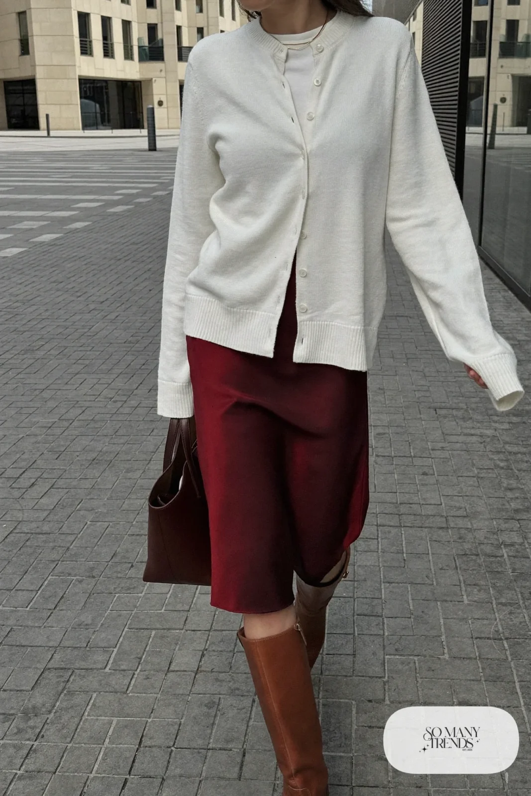 Woman in a burgundy satin midi skirt, cream knit cardigan, tall brown boots and a gold necklace, outfit 14