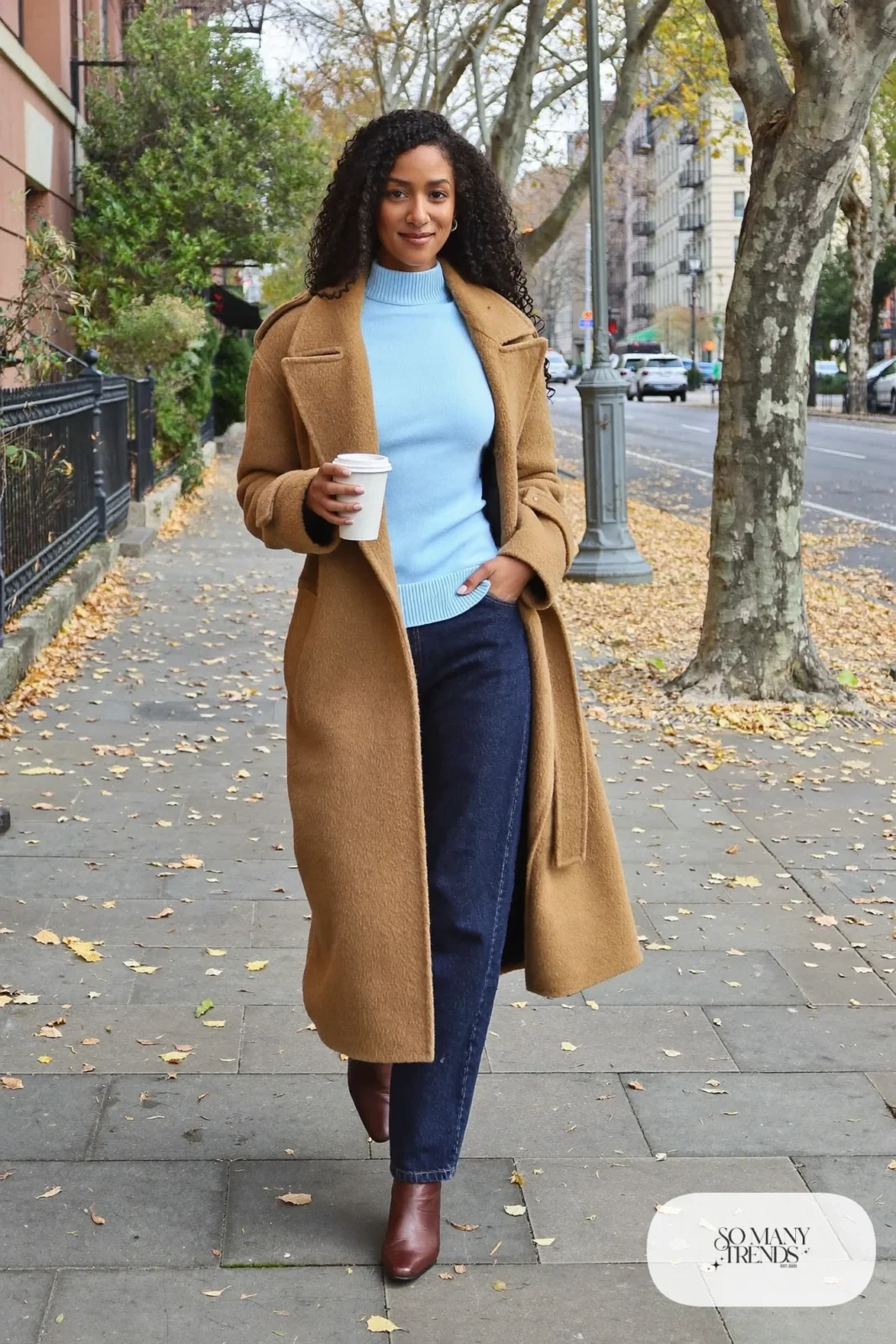 Woman with a straight athletic figure in a long camel wrap coat belted at the waist, a baby blue knit sweater, straight dark jeans and brown leather ankle boots, outfit 1