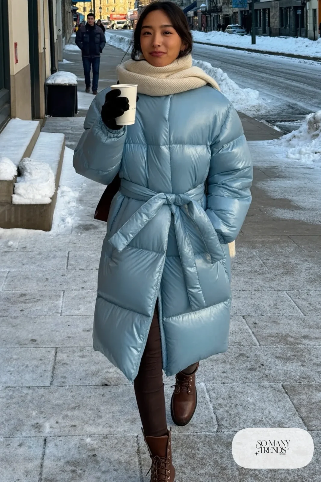 Woman with a straight athletic figure in a long baby blue belted puffer coat, slim dark pants and brown boots on a winter street, outfit 14