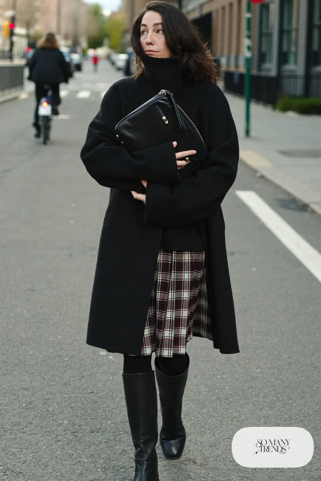 Woman in a black funnel neck coat over a burgundy plaid pleated midi skirt, black knit top, black tights and tall black boots, outfit 7