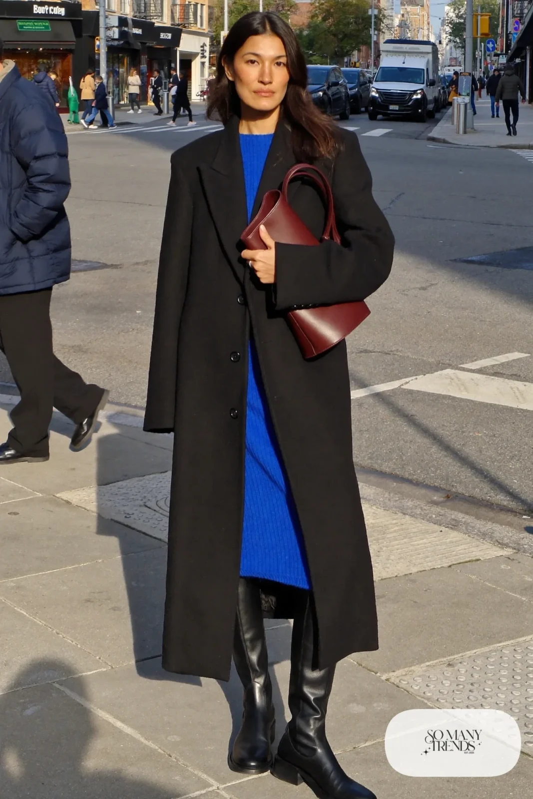 Woman in a long black wool coat over a cobalt blue knit midi dress with a wine leather bag and tall black boots, outfit 30