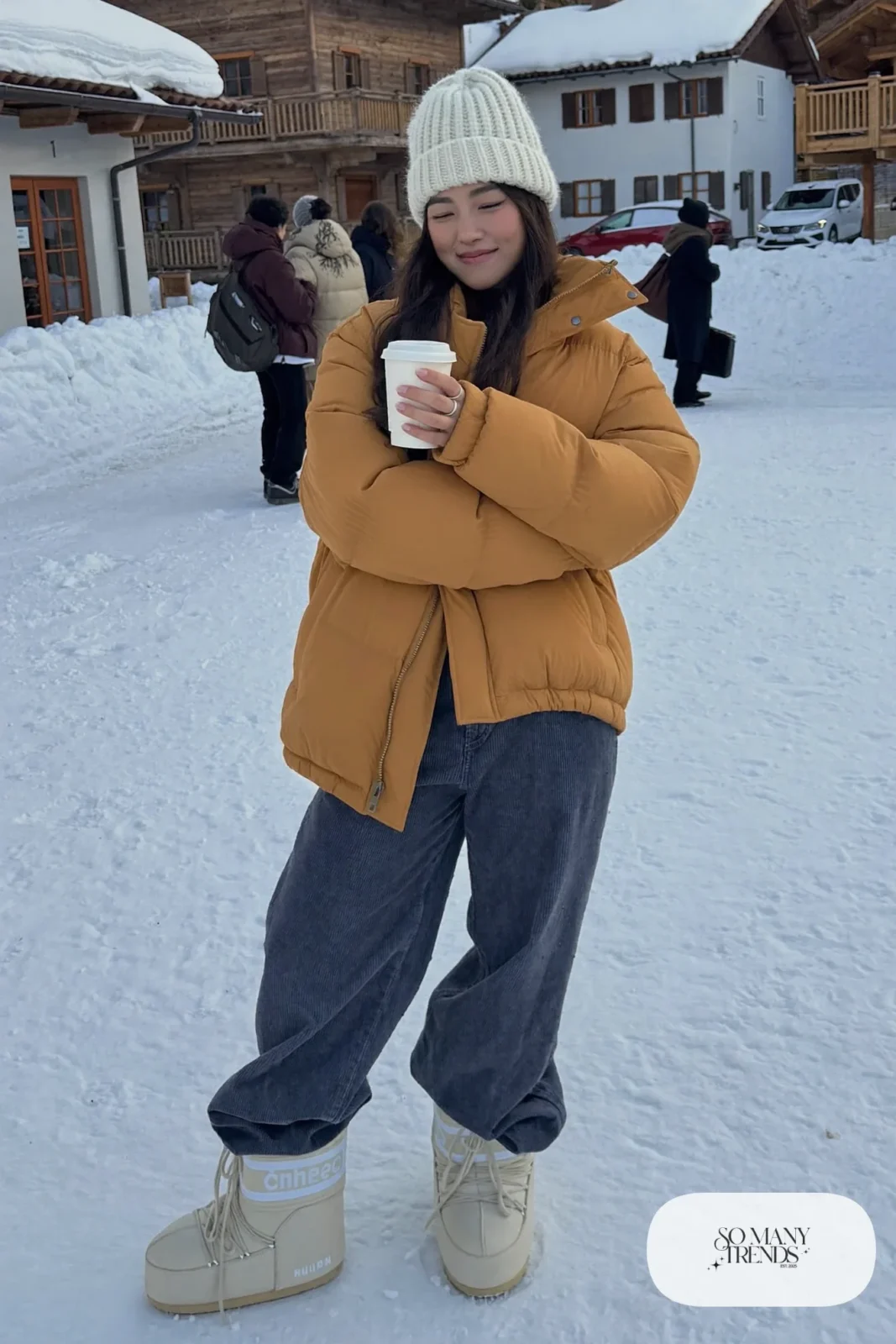 Woman in terracotta long puffer jacket, wide-leg indigo corduroy pants, cream moon boots and a cream beanie in the snow, outfit 1