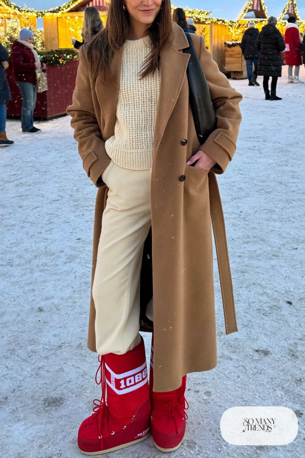 Woman in long camel belted coat, cream straight pants, red moon boots and a cream knit sweater in a snowy square, outfit 9