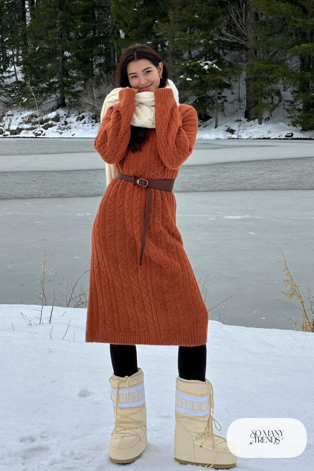 Woman in terracotta cable-knit midi sweater dress, black fleece tights, cream moon boots and a belt in the snow, outfit 12