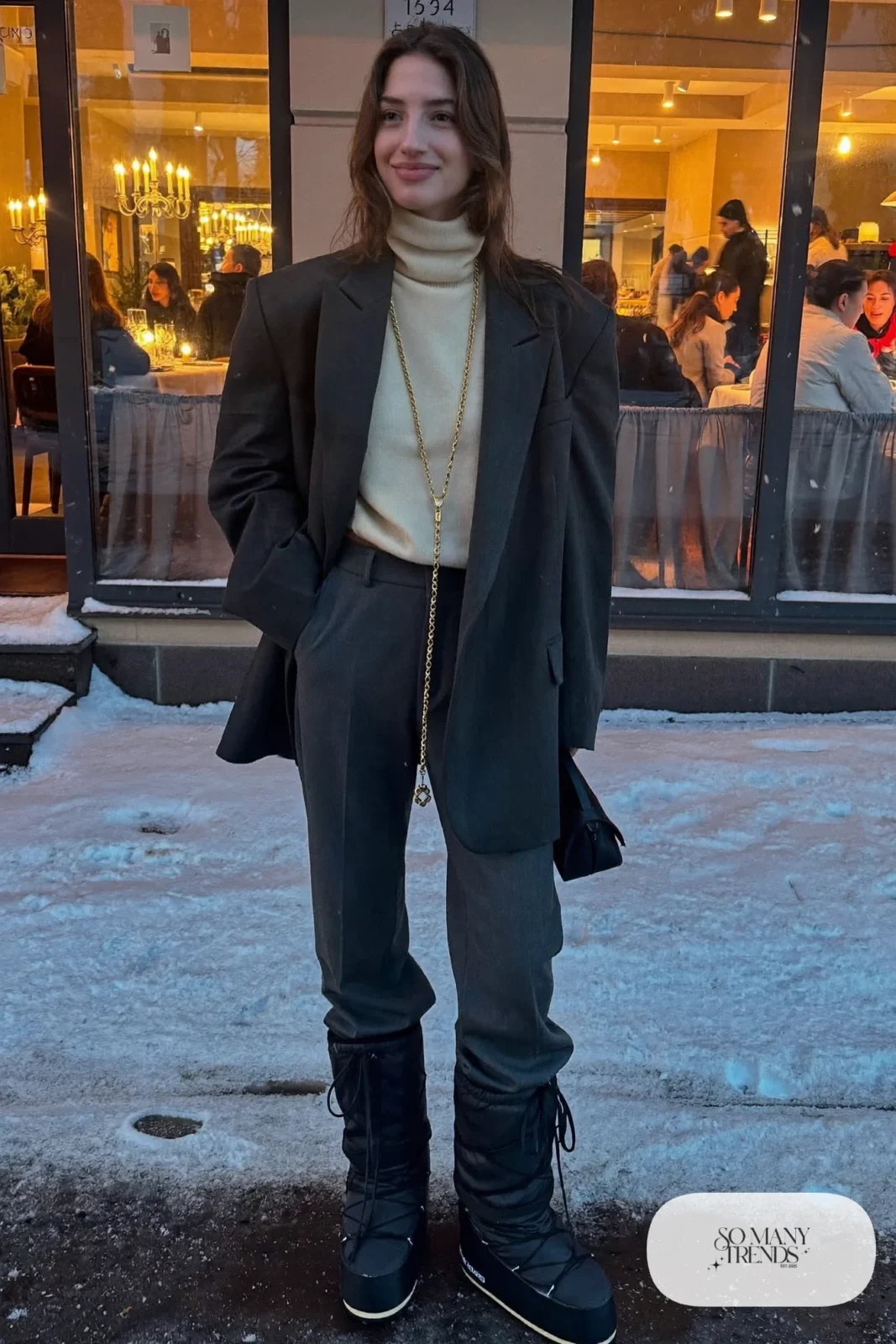 Woman in charcoal oversized blazer, cream turtleneck, straight charcoal wool pants and black moon boots in the snow, outfit 14