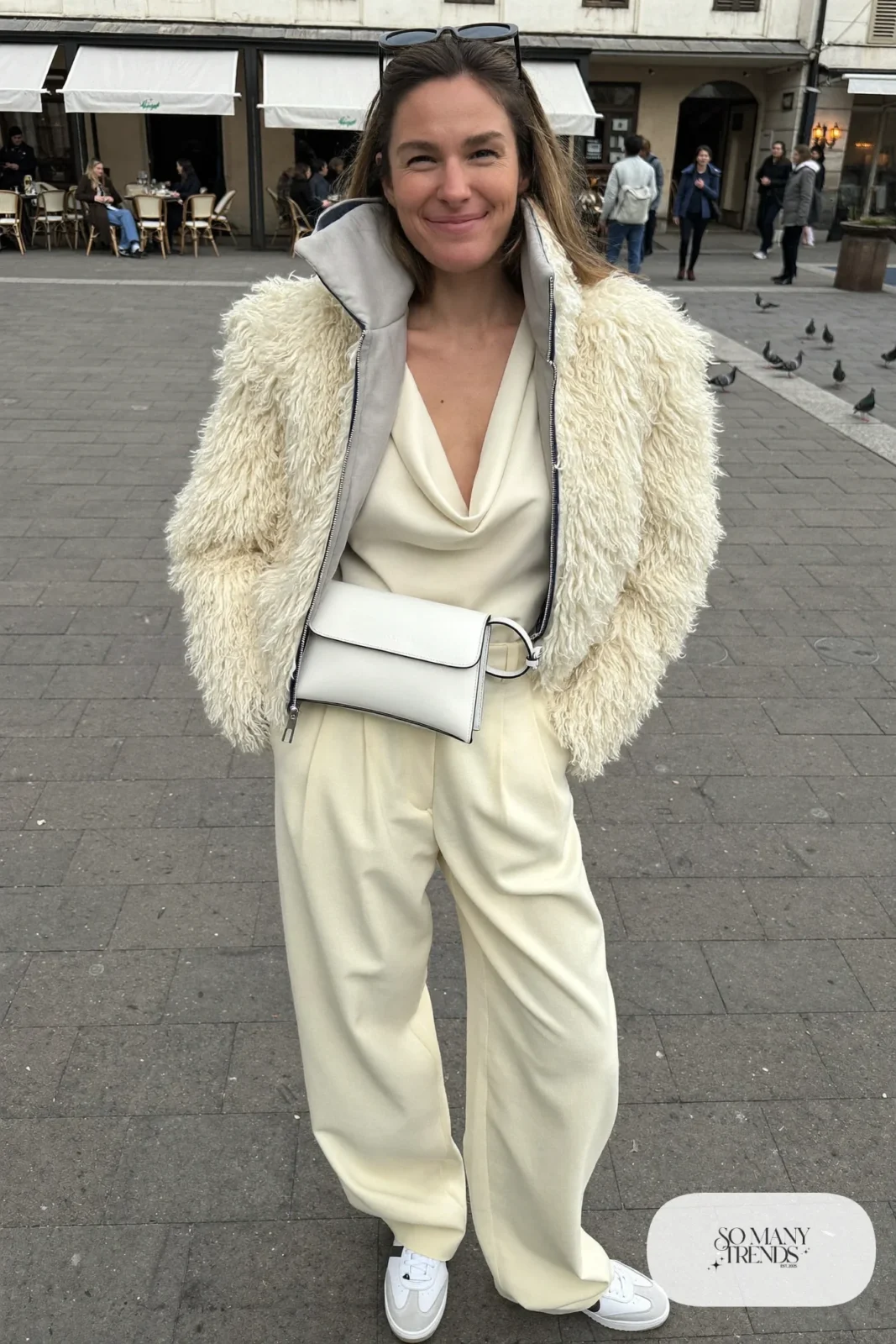 Woman in ivory shearling short jacket, cream draped top, cream wide pleated pants, white sneakers and a white structured bag, outfit 28