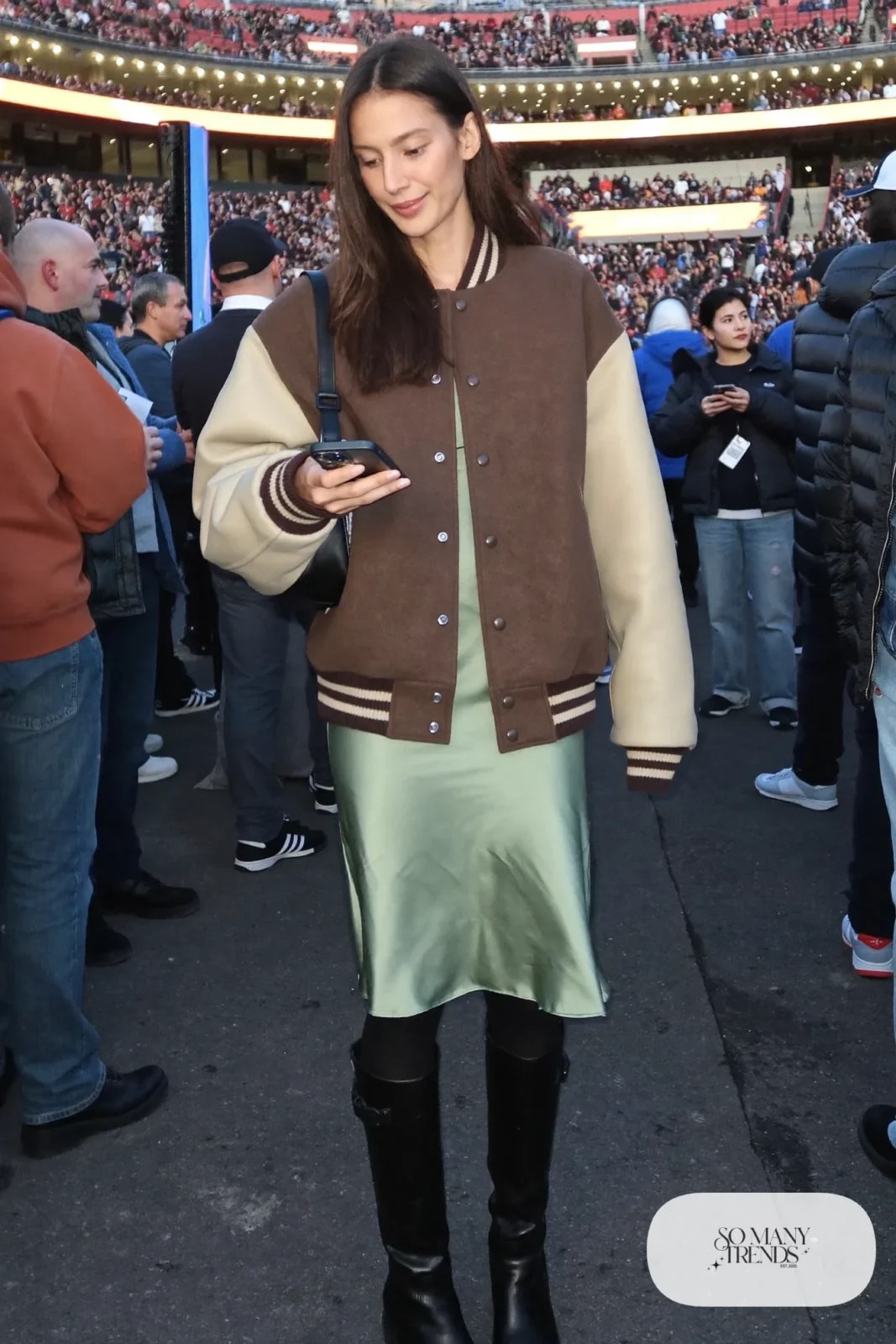 Woman in an oatmeal and espresso varsity jacket, sage satin slip skirt, black tights and tall black boots, basketball game outfit 6