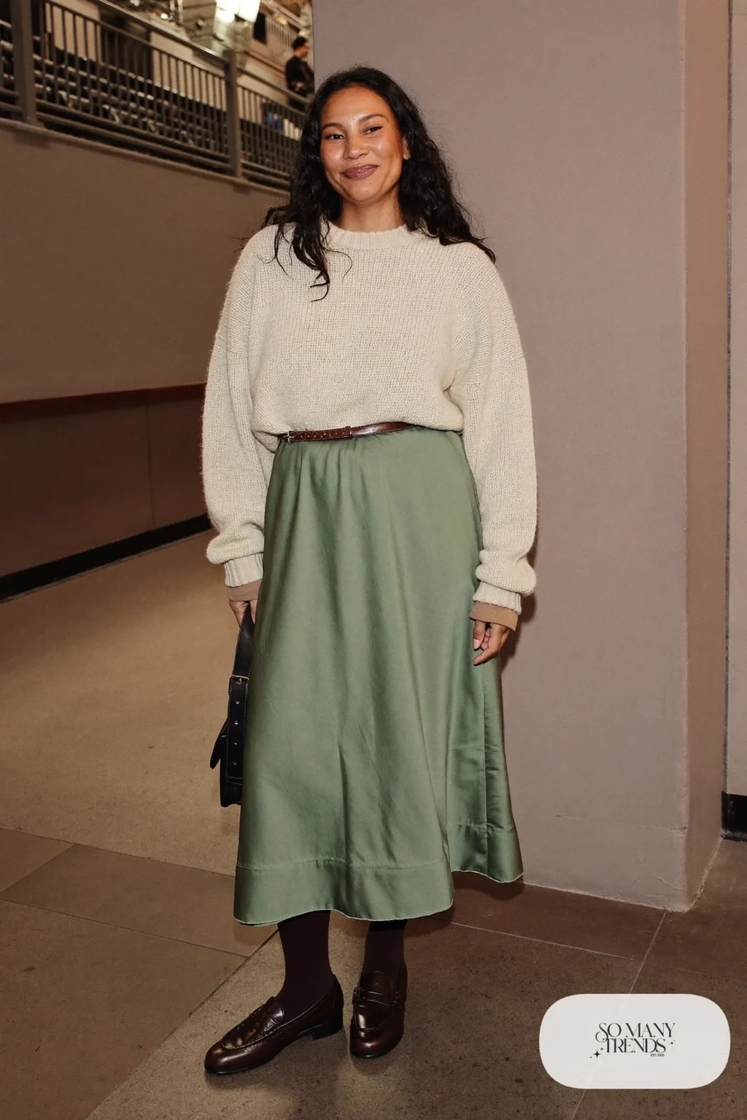 Woman in a sage satin midi skirt, chunky oatmeal knit, opaque tights and brown loafers, basketball game outfit 13