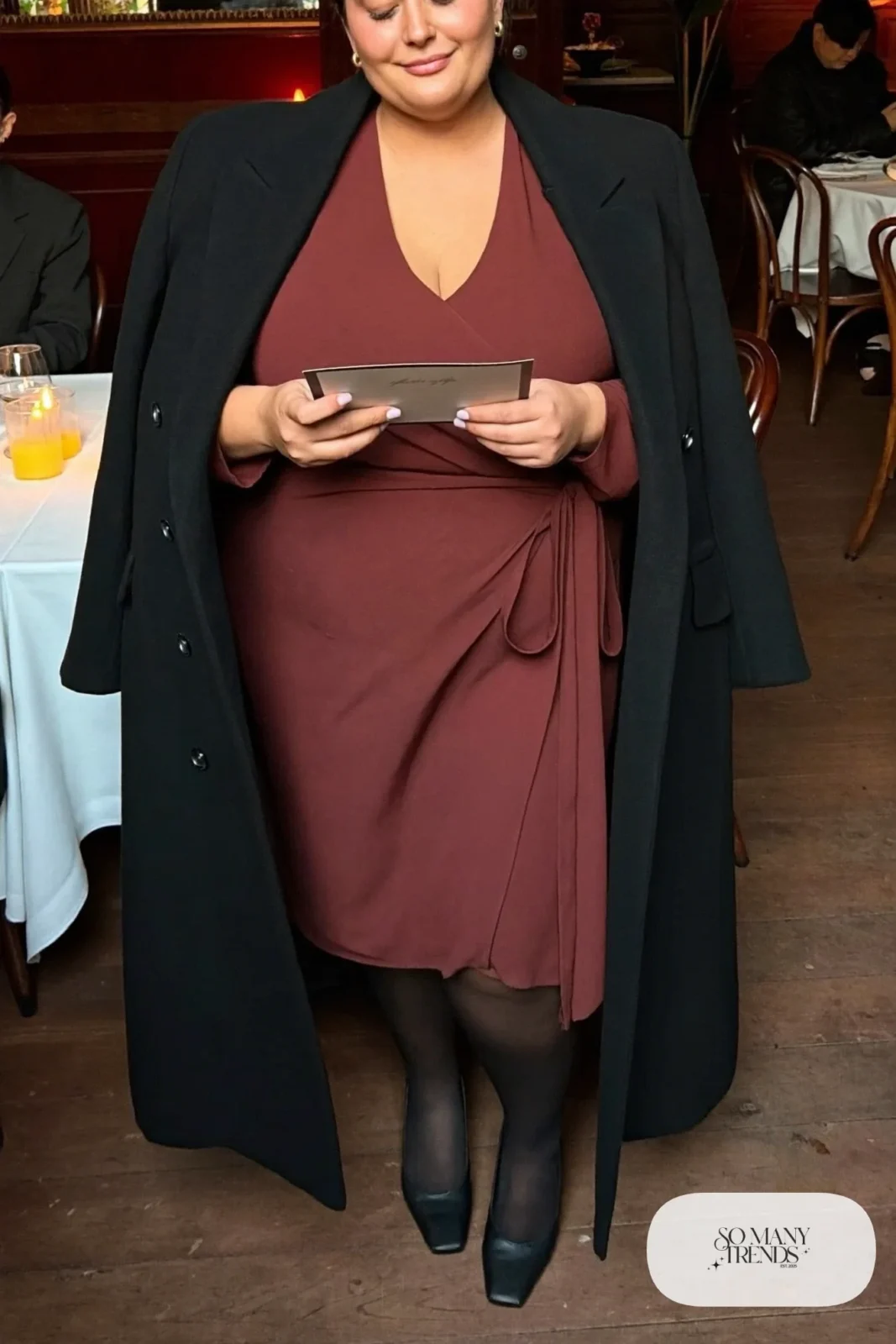 Plus size woman in oxblood wrap midi dress with black tights, black pumps and black long coat, outfit 1