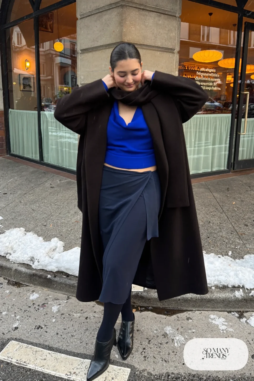 Plus size woman in cobalt cowl top, navy wrap midi skirt, black long coat and black ankle boots, outfit 12