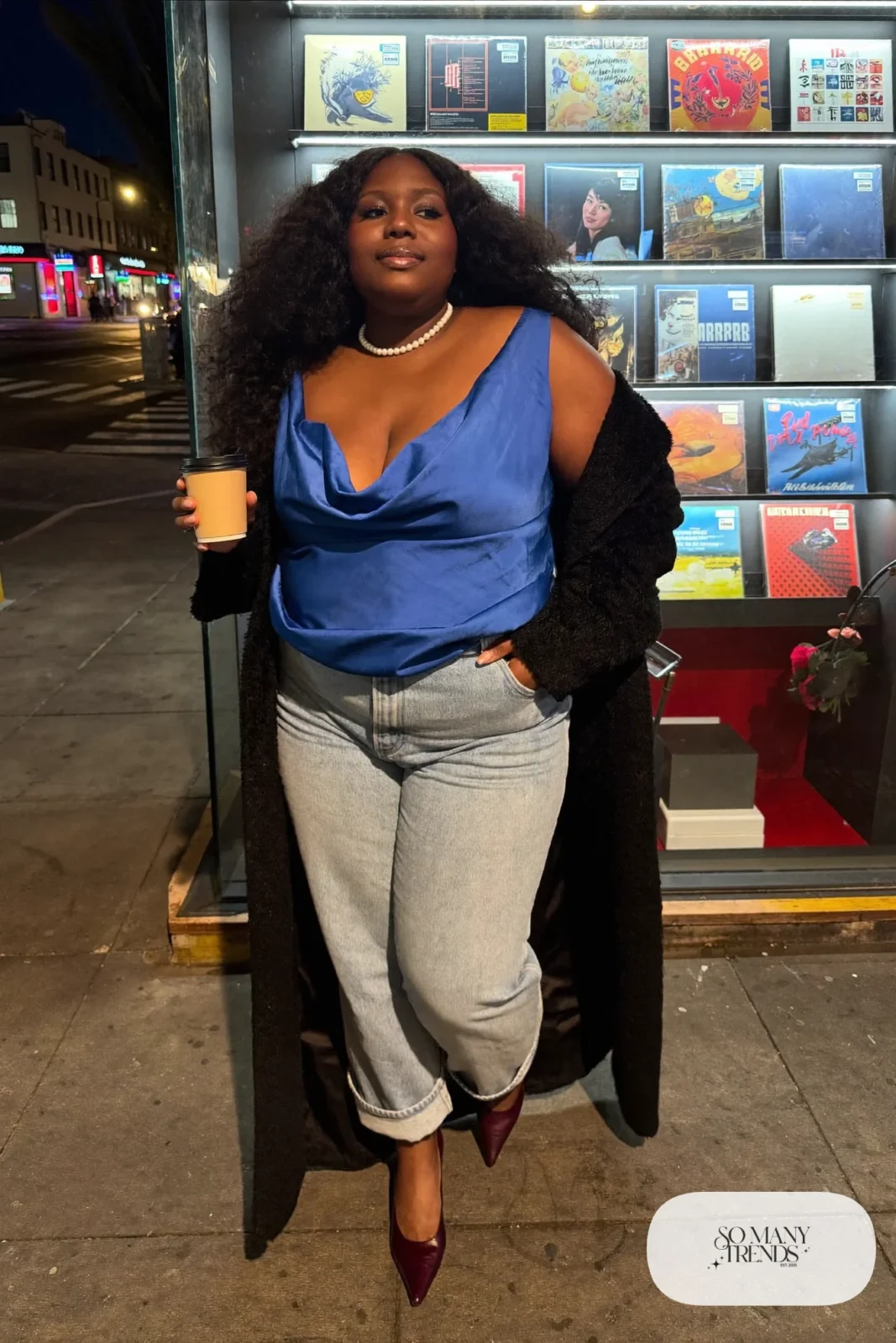 Plus size woman in light barrel jeans, cobalt satin cowl top, black long coat and oxblood pointed booties, outfit 14