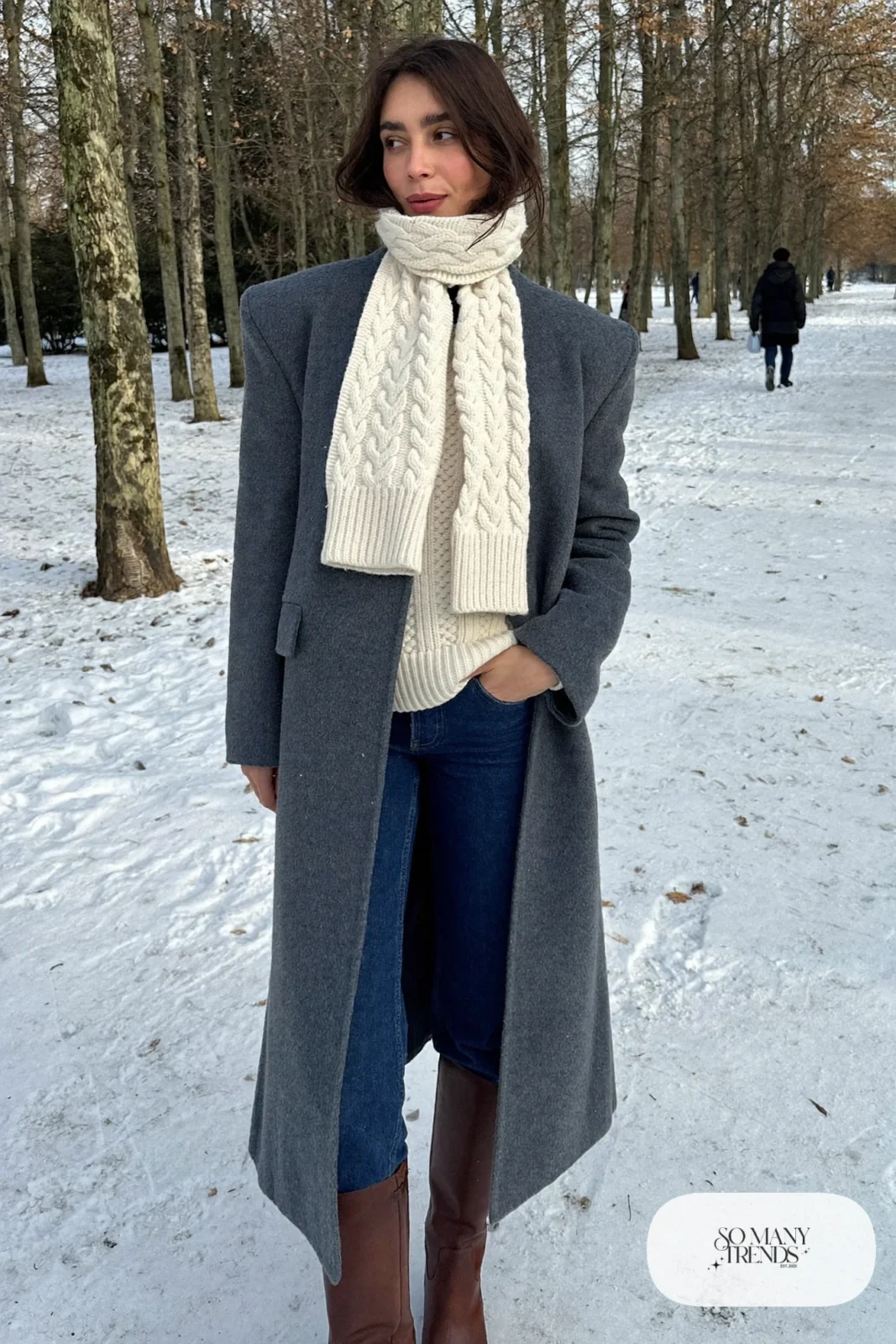 Woman in a slate blue knee-length wool coat, cream knit sweater, dark straight jeans, cream scarf and brown tall boots in snow, outfit 1
