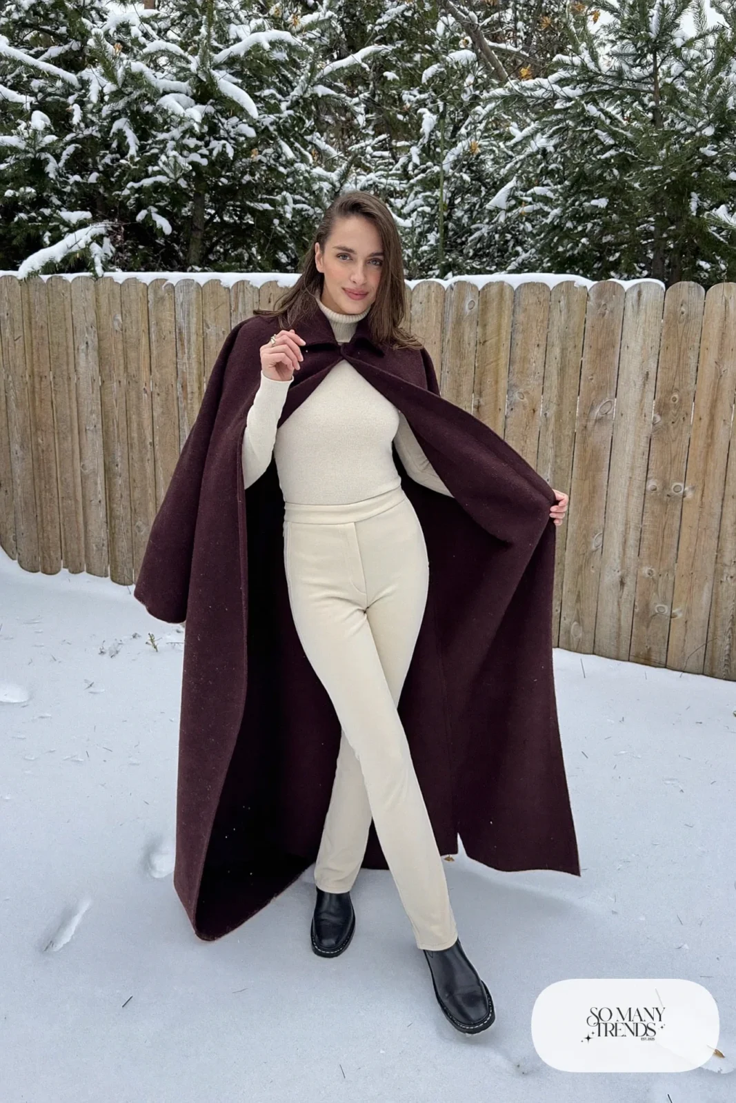 Woman in a wine wool cape, cream turtleneck, cream slim pants and black ankle boots in a snowy yard, outfit 2
