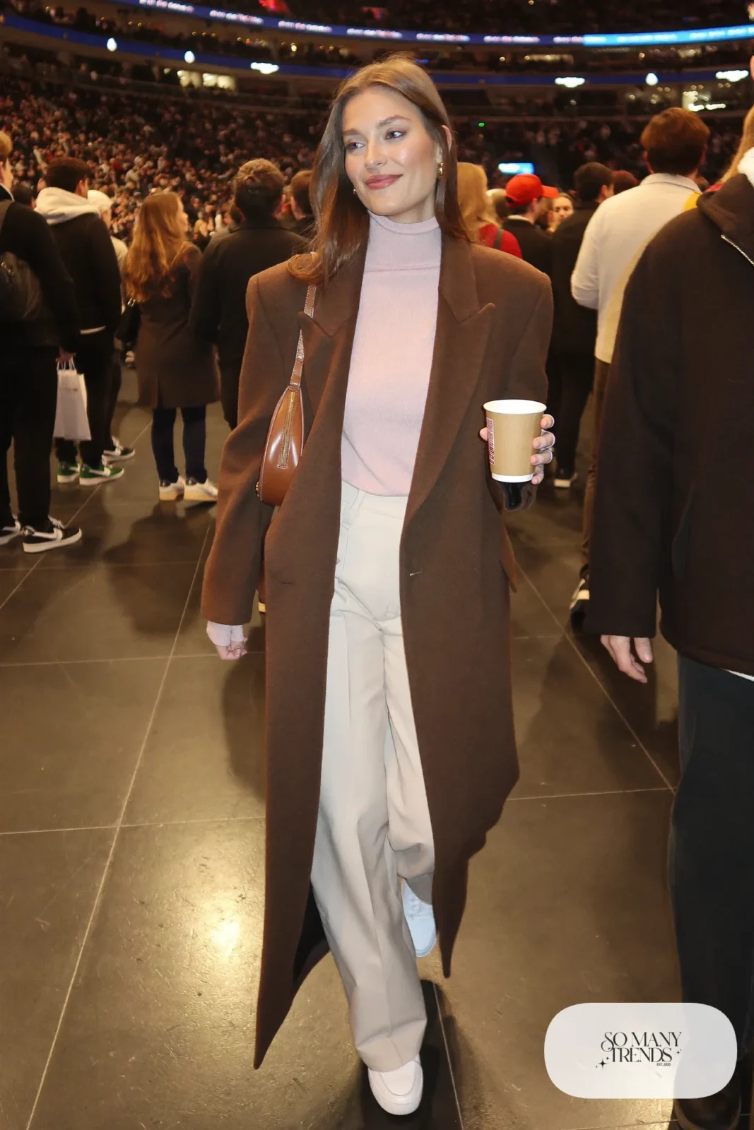Woman in long espresso brown wool coat, pale pink ribbed turtleneck, cream wide-leg pants and white leather sneakers, hockey game outfit 1