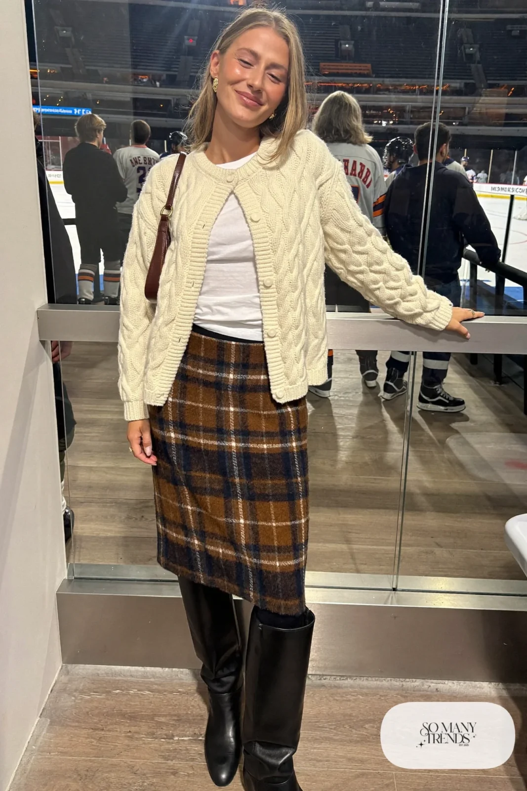 Woman in cream cable knit cardigan, brown plaid wool midi skirt, black tights and black knee-high boots, hockey game outfit 18