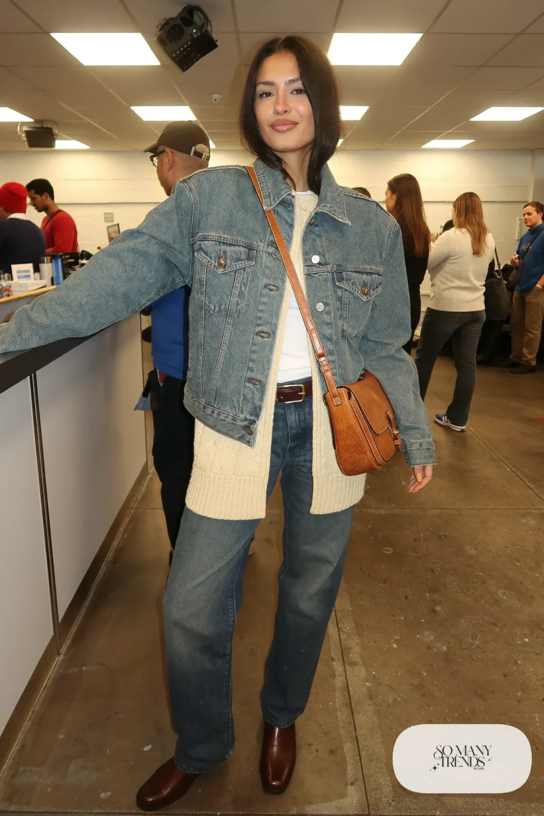 Woman in a washed denim jacket, cream knit vest, dark jeans, leather belt and brown boots, concert outfit 3
