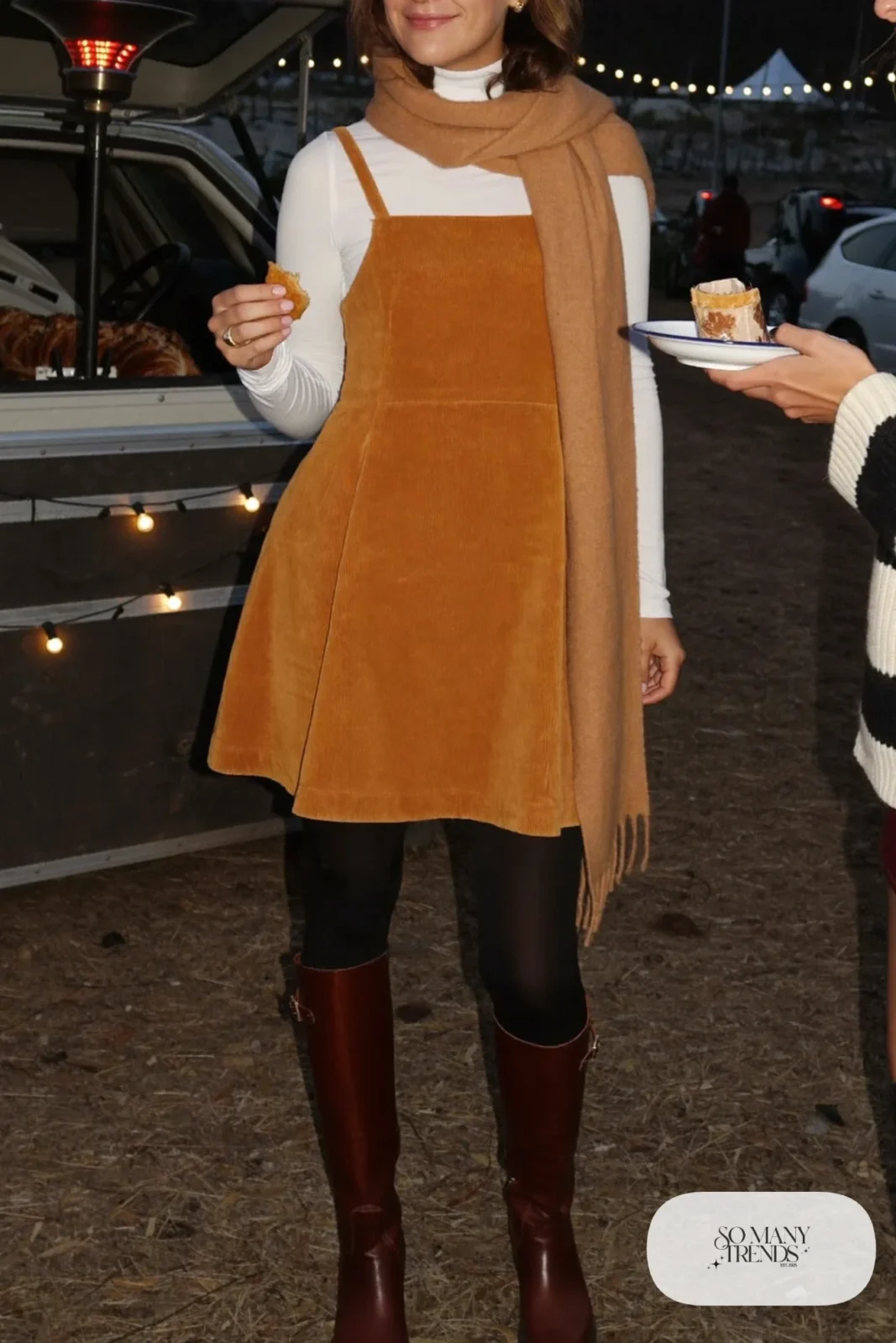 Woman in a mustard corduroy pinafore dress, white turtleneck, tights and brown tall boots, concert outfit 9