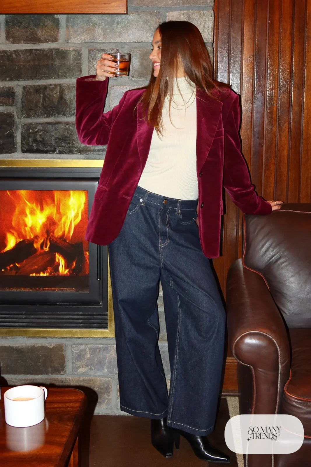 Woman in a burgundy velvet blazer, cream ribbed turtleneck, wide-leg dark indigo jeans and black ankle boots near a lodge fireplace, outfit 3