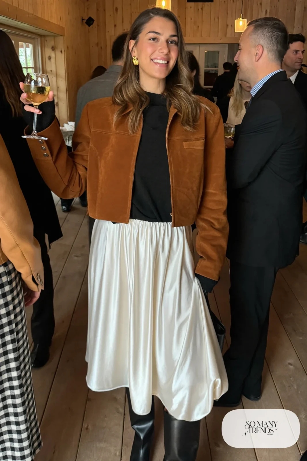 Woman in a cognac cropped suede jacket, black cashmere sweater, pearl satin midi skirt, black opaque tights and tall black boots at a lodge party, outfit 5