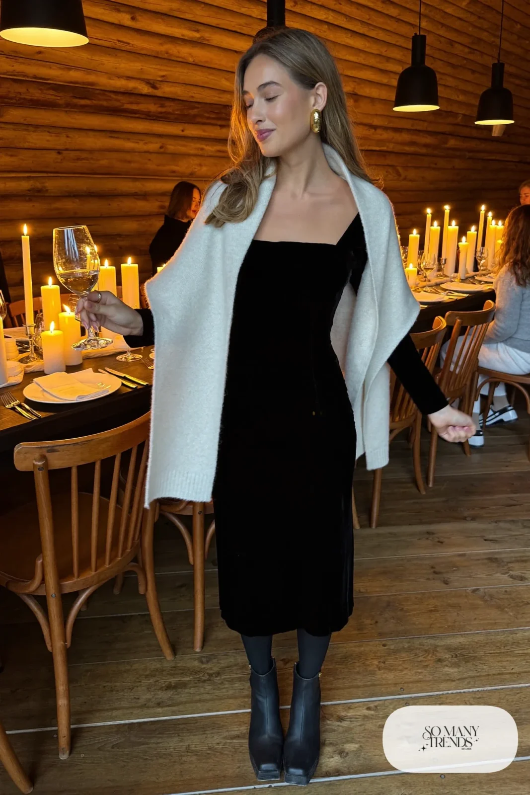 Woman in a black long-sleeve velvet midi dress with a pearl wool wrap over her shoulders, sheer black tights and black block-heel ankle boots at a candlelit lodge dinner, outfit 9