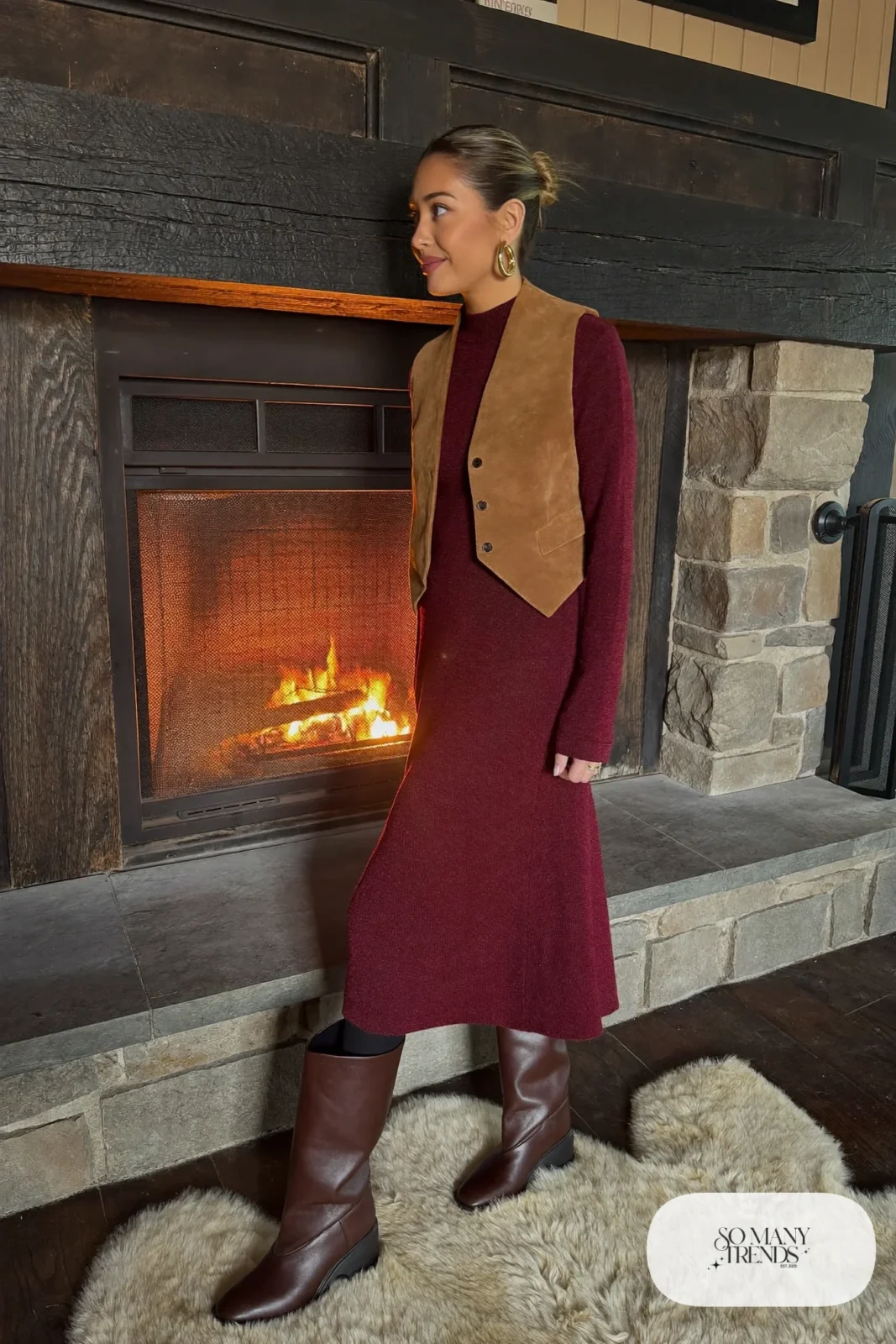 Woman in a burgundy knit midi dress with a camel suede vest, thick black tights and tall brown boots by a lodge fire, outfit 10