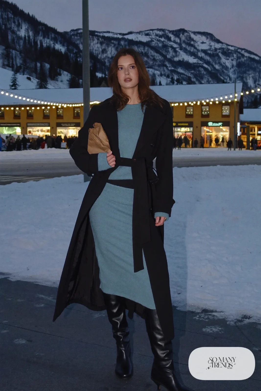 Woman in a black belted wool trench coat over an icy blue knit sweater and midi skirt with tall black boots on a snowy mountain street, outfit 11