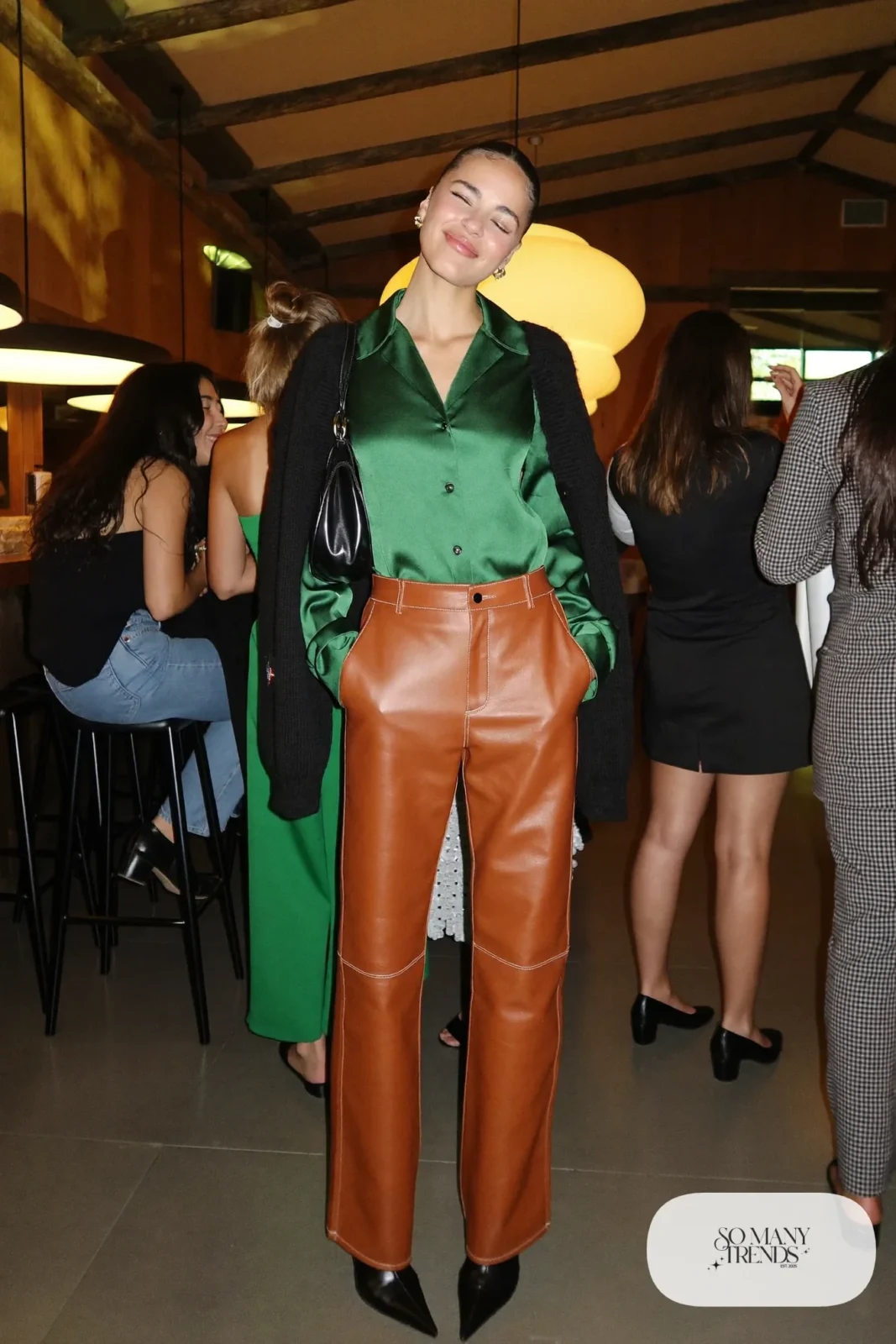 Woman in an emerald satin blouse tucked into cognac faux leather pants with a black cashmere cardigan over her shoulders and black pointed ankle boots at a lodge party, outfit 13