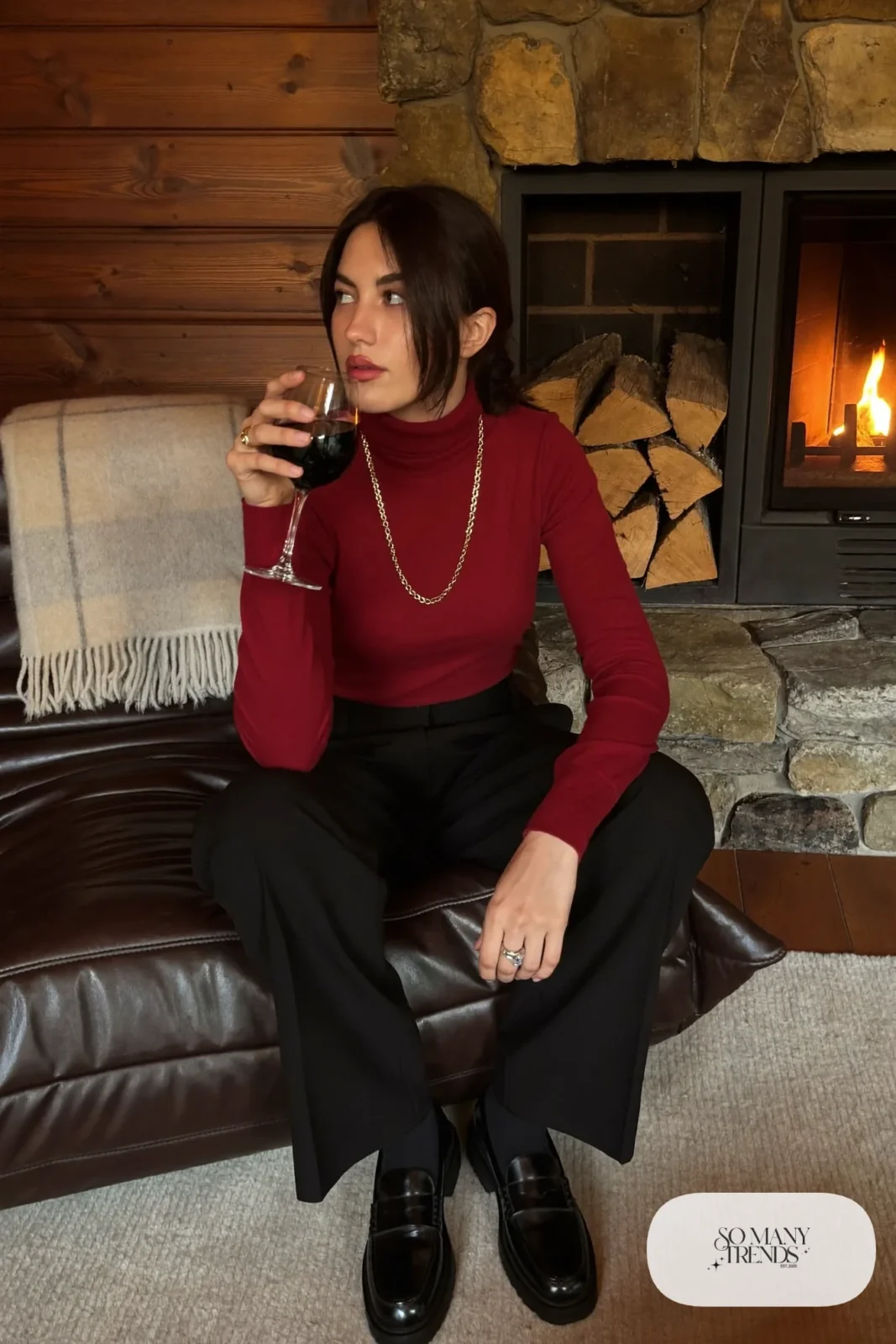 Woman in a burgundy merino turtleneck, black high-waisted tailored pants, gold chain necklace and black lug-sole loafers at a lodge fireside, outfit 15