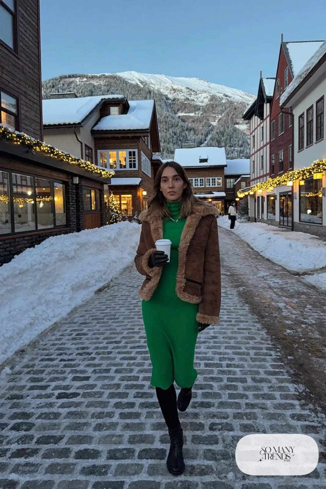 Woman in a cognac cropped shearling jacket over an emerald knit midi dress with black opaque tights and black ankle boots in a mountain town, outfit 16