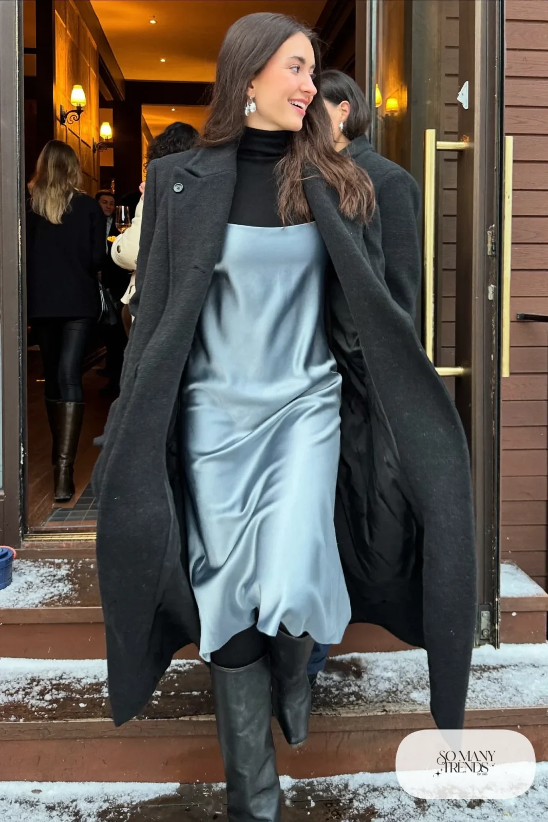 Woman in an icy blue satin slip dress layered over a black turtleneck with a long black wool coat, opaque tights and tall black boots arriving at a lodge party, outfit 17