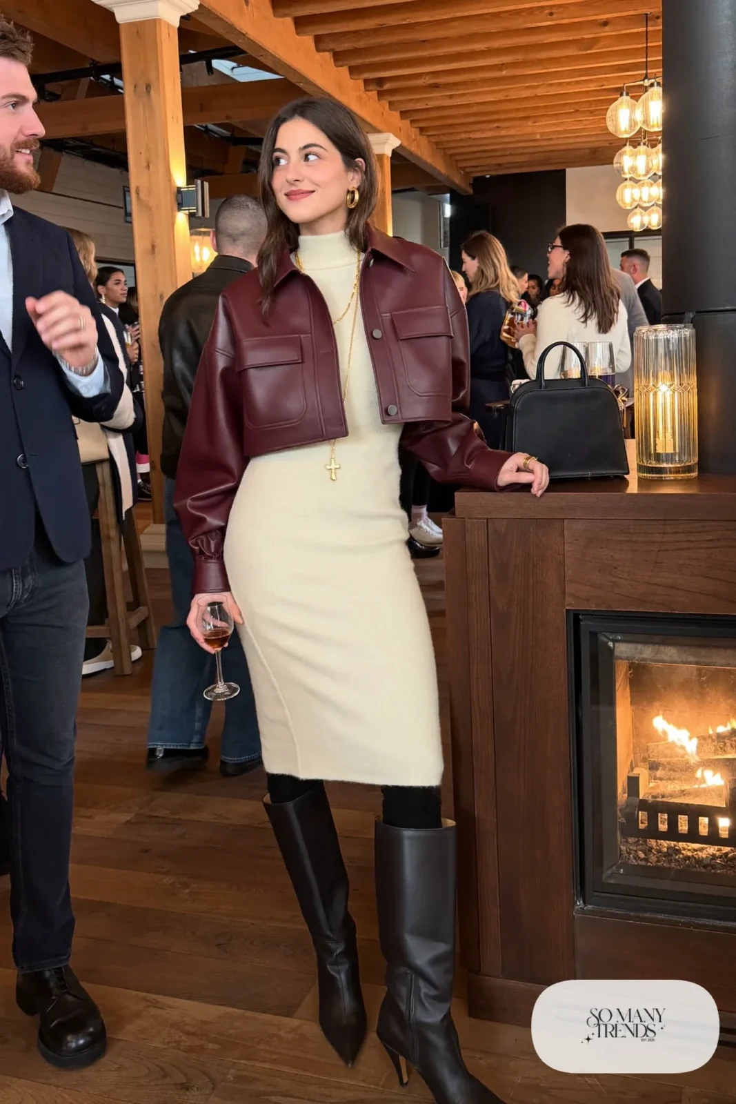 Woman in a burgundy cropped leather jacket over a cream knit midi dress with opaque black tights and tall black boots at a lodge party, outfit 25