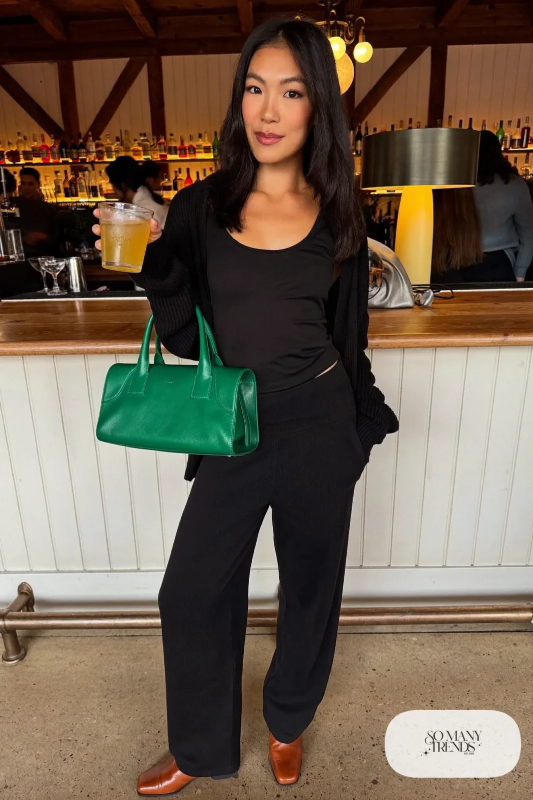 Woman in a black knit ribbed cardigan and wide pants co-ord with cognac boots and an emerald leather bag at a lodge bar, outfit 28
