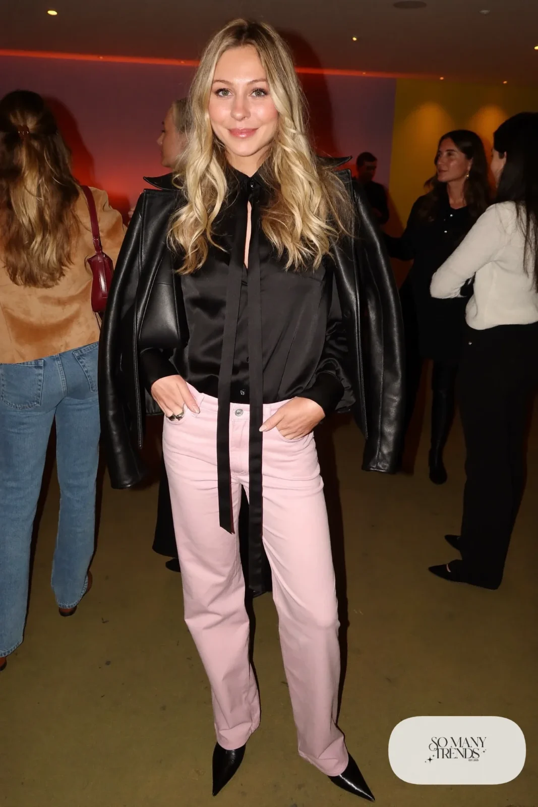 Woman in a black tie-neck blouse, pale pink straight pants, black leather jacket and pointed black boots, outfit 23