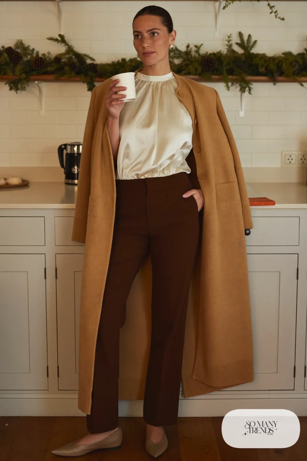 Woman in a long camel cardigan, cream silk-feel shell, dark brown straight pants and pointed flats in a kitchen, outfit 2