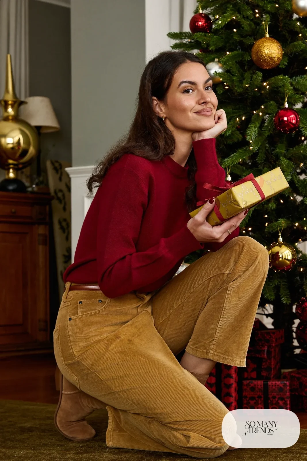 Woman in a deep red fine-knit sweater, sand corduroy straight pants and tan suede ankle boots at a living room tree, outfit 3
