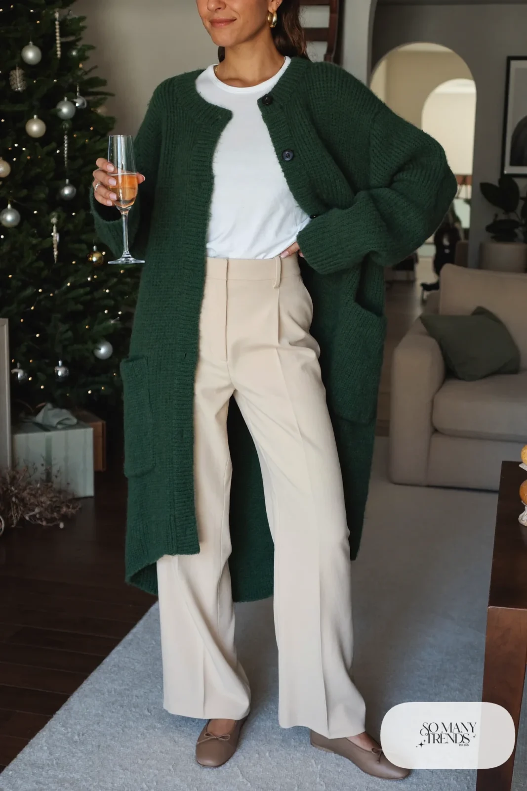 Woman in a forest green cardigan, white tee, cream wide-leg pants and taupe flats near a decorated tree, outfit 6