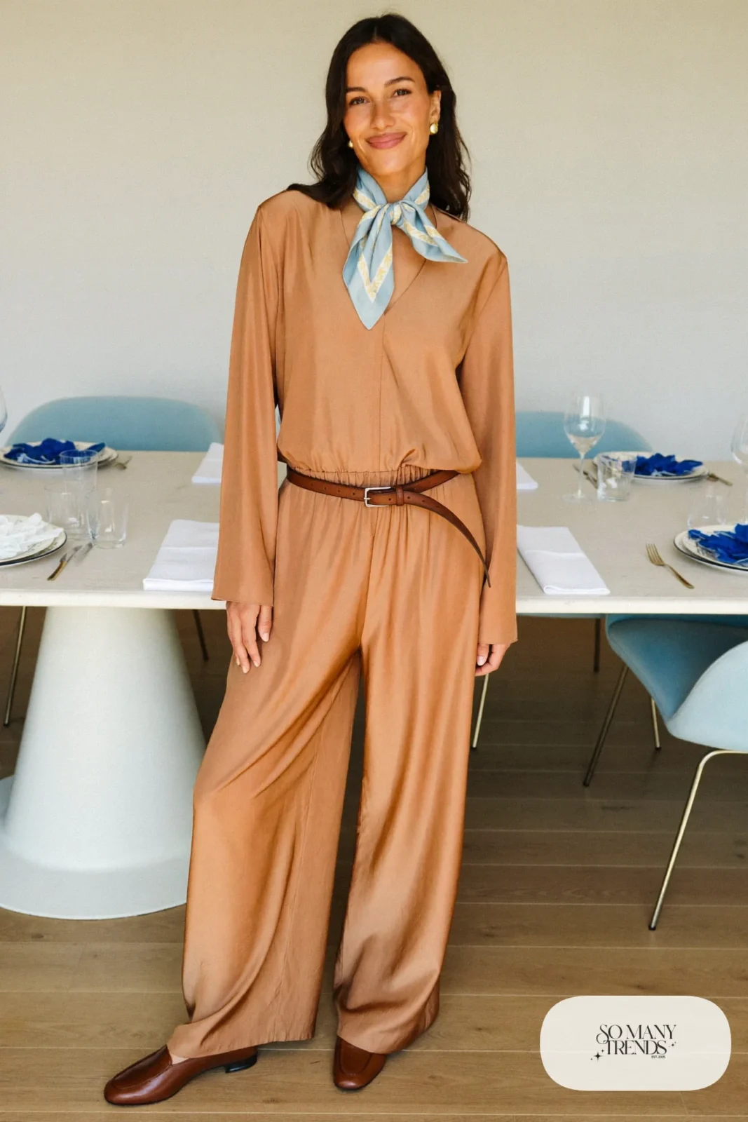 Woman in a sand wide-leg jumpsuit with a thin belt, baby blue scarf and brown loafers in a bright dining room, outfit 15
