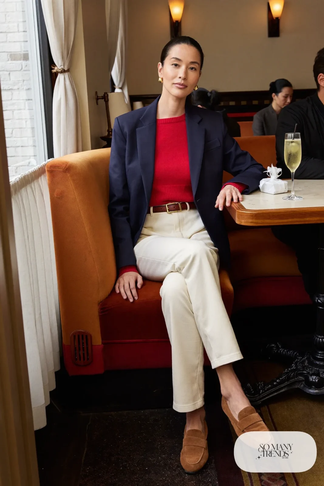 Woman in a navy blazer, wine red knit, cream pants and brown suede loafers at a restaurant table, outfit 16