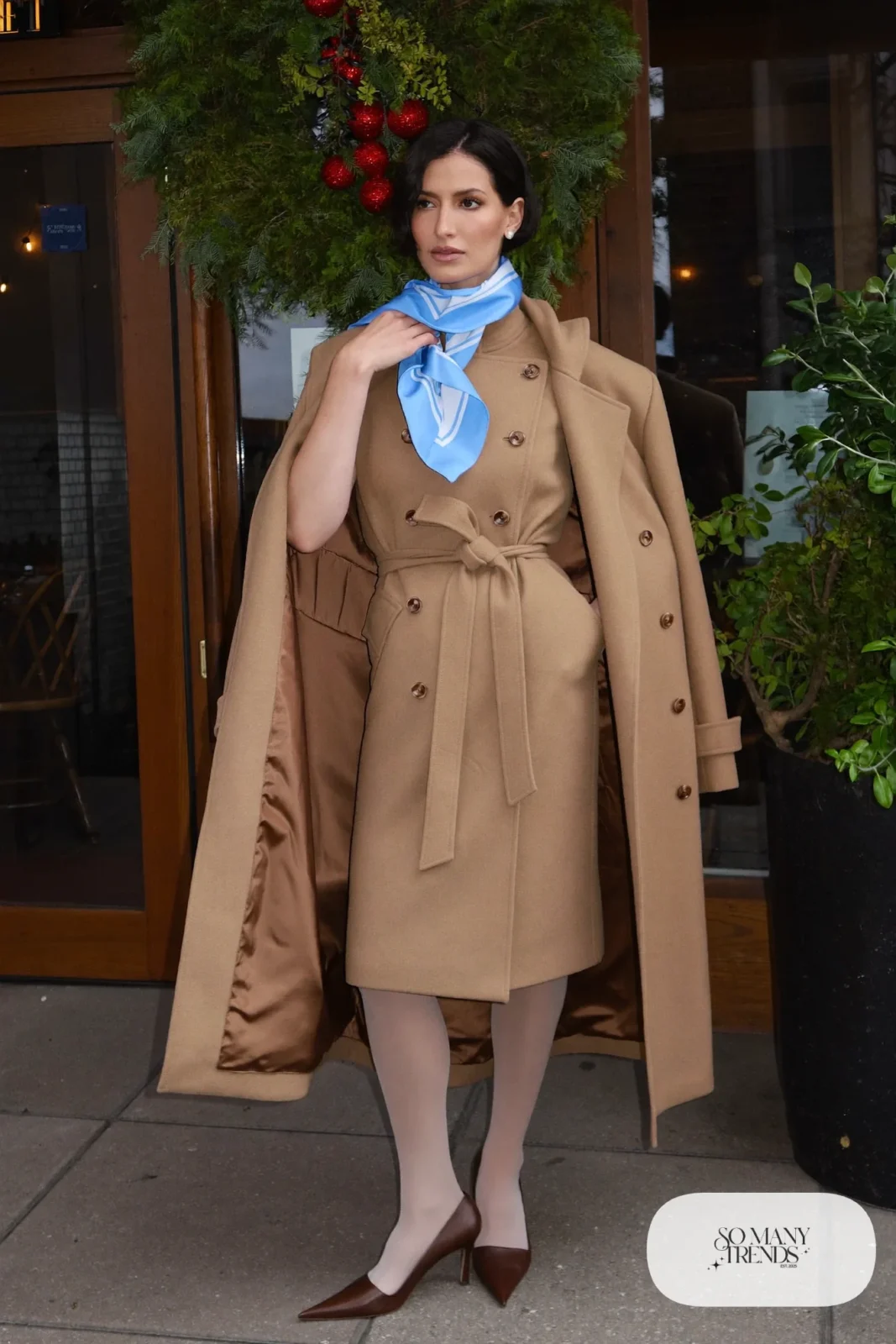 Woman in a belted camel wool coat dress with a baby blue scarf, sheer tights and brown pumps at a restaurant entrance, outfit 17