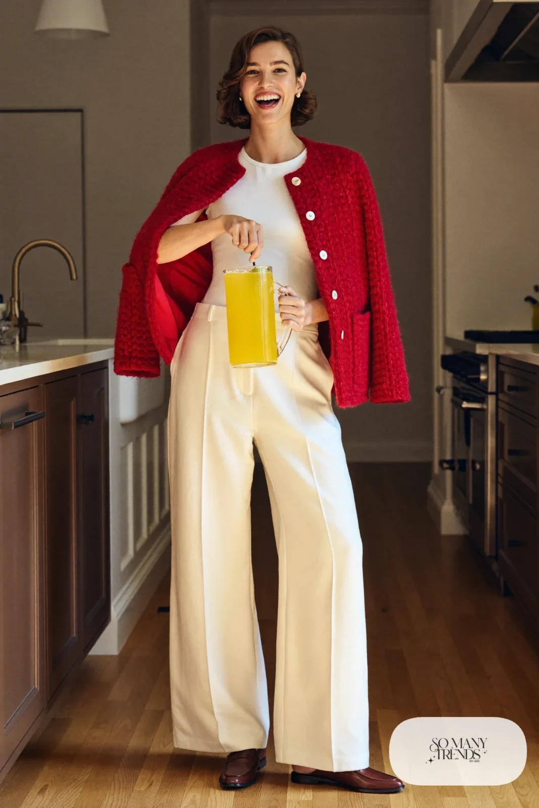 Woman in a chunky red cardigan, white knit tee, cream wide-leg pants and brown loafers in a warm kitchen, outfit 27