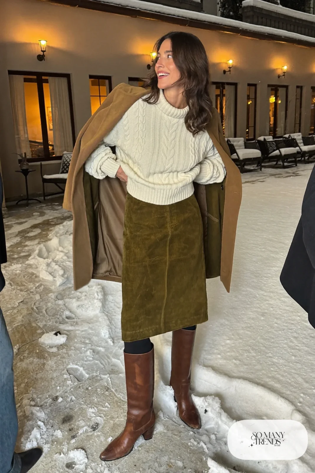 Woman in a moss green suede midi skirt, cream cable knit sweater, black opaque tights and tall brown leather boots, outfit 12