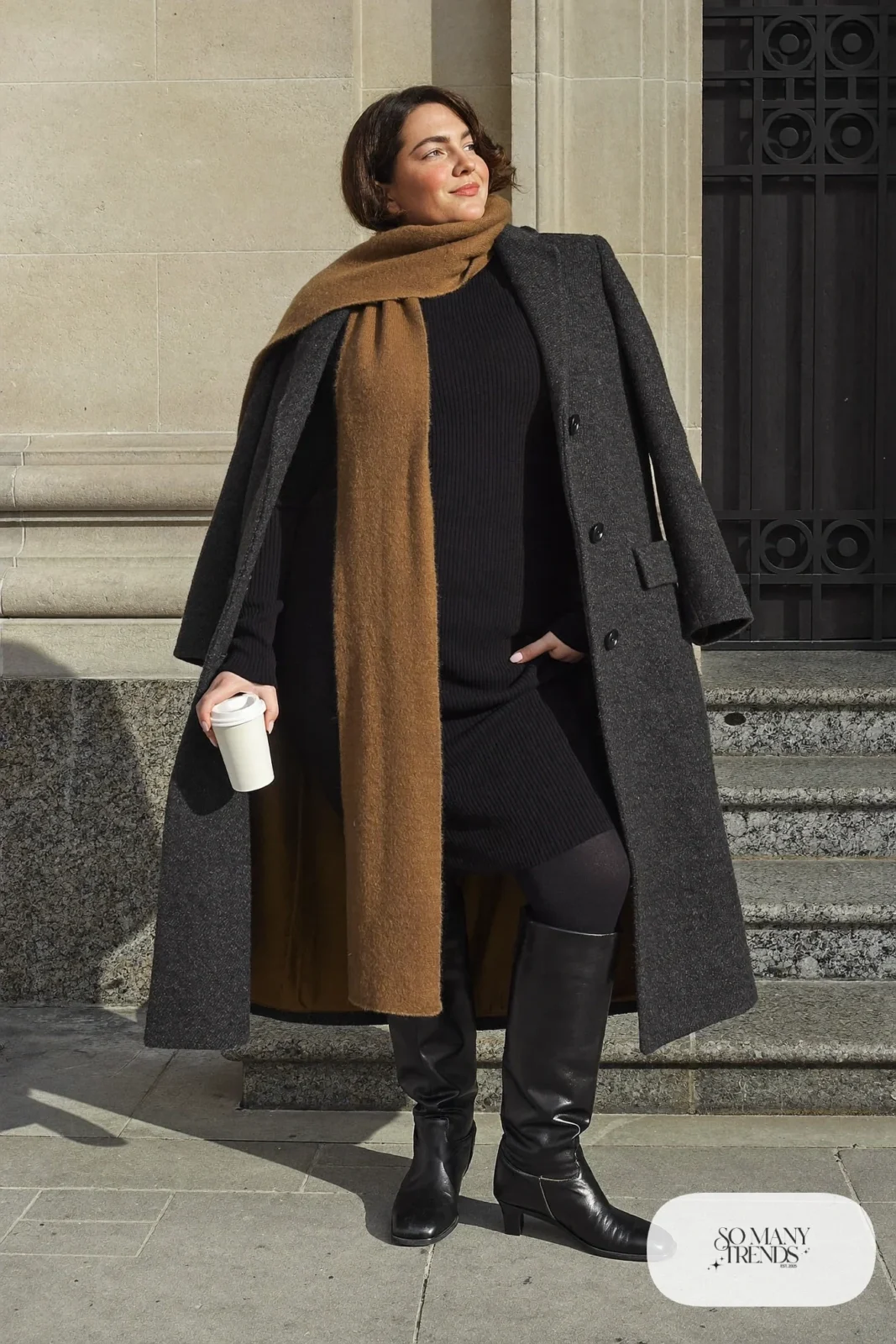Woman in a black long-sleeve ribbed knit midi dress, black opaque tights, tall black leather boots and a camel scarf, outfit 3