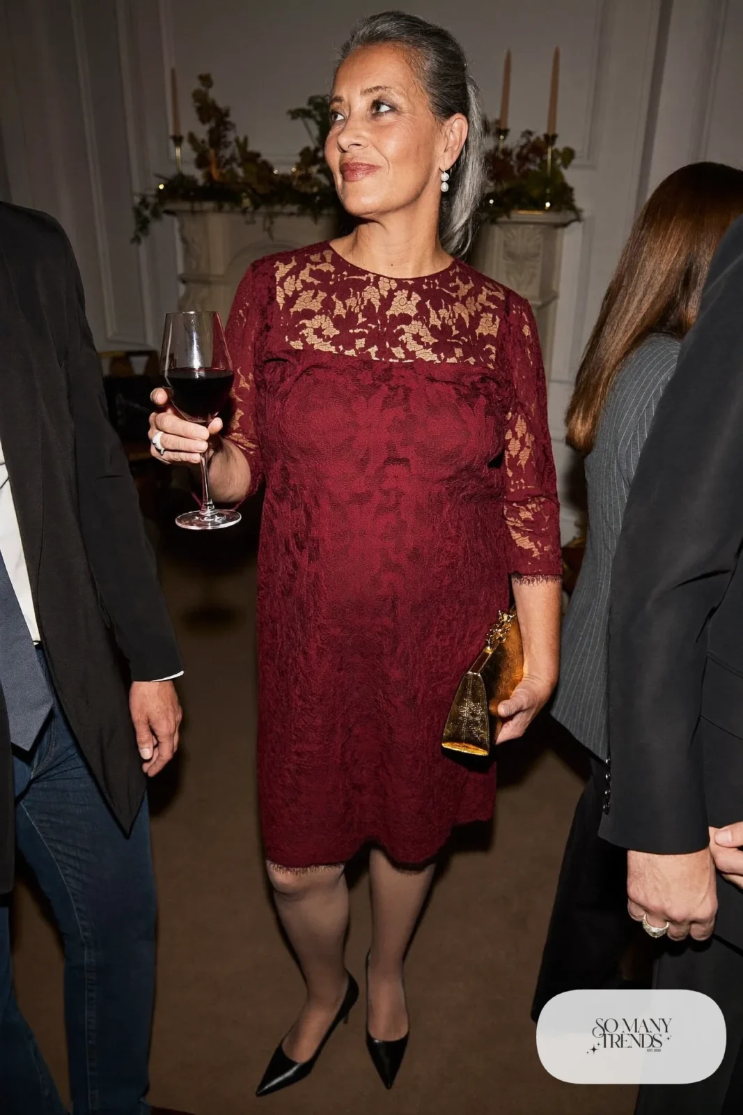 Woman in a burgundy lace knee-length sheath dress with sleeves, black pumps and a gold clutch, outfit 5