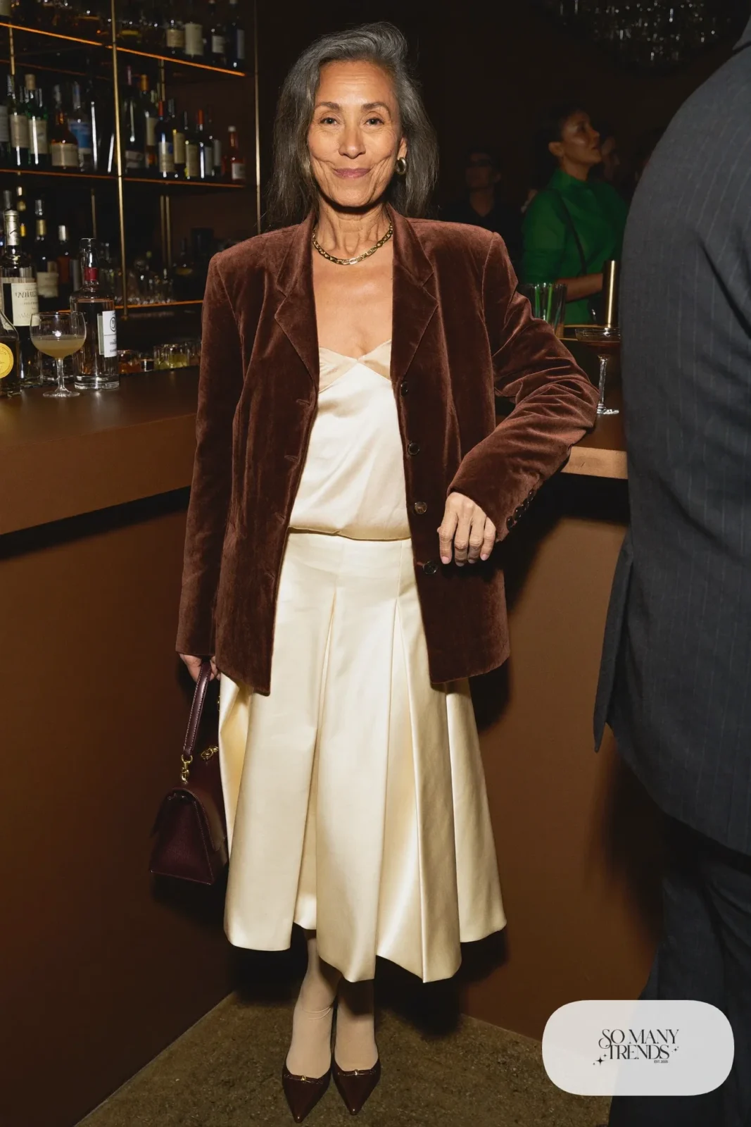 Woman in a chocolate brown velvet blazer, cream pleated midi skirt, sheer nude tights and brown pointed pumps, outfit 8