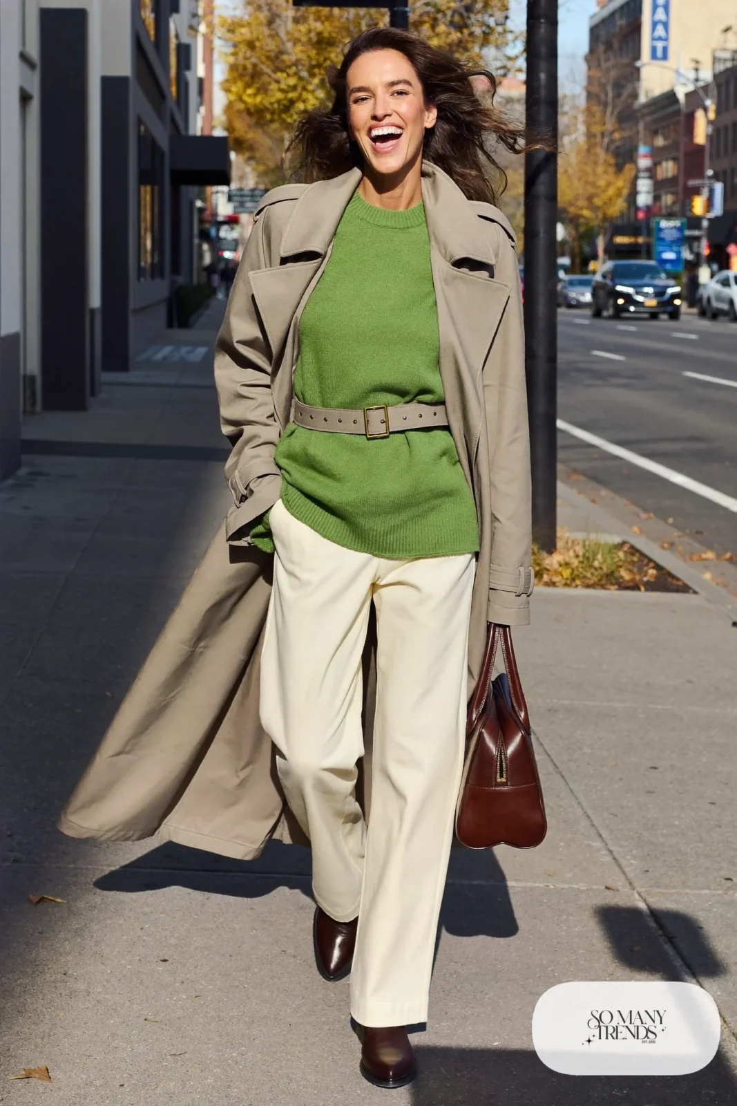 Woman in belted stone trench coat, pine green knit sweater, cream pants and brown boots, outfit 9