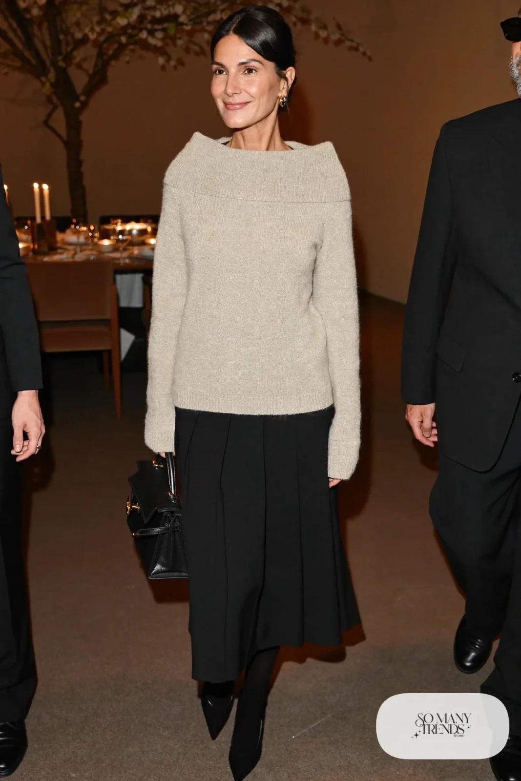 Woman in an oatmeal fine knit top with a black pleated midi skirt, sheer tights and black pointed pumps, photo 13